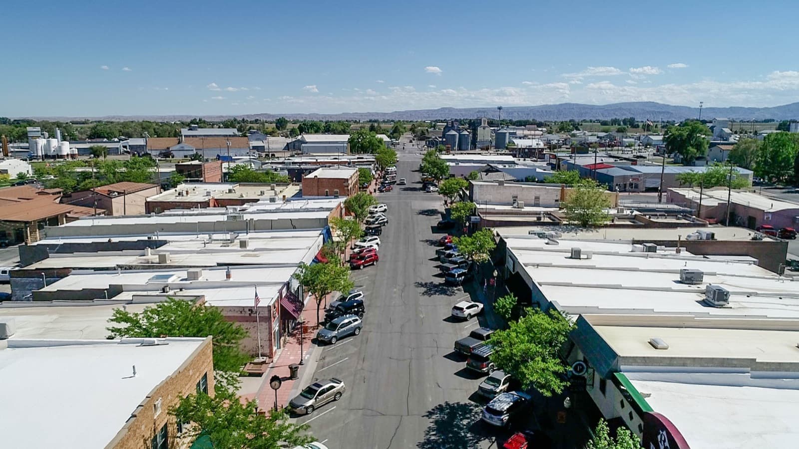 Aerial view down Powell's downtown main street, Wyoming — brick storefronts and grain elevators with farmland stretching to the distant mountains