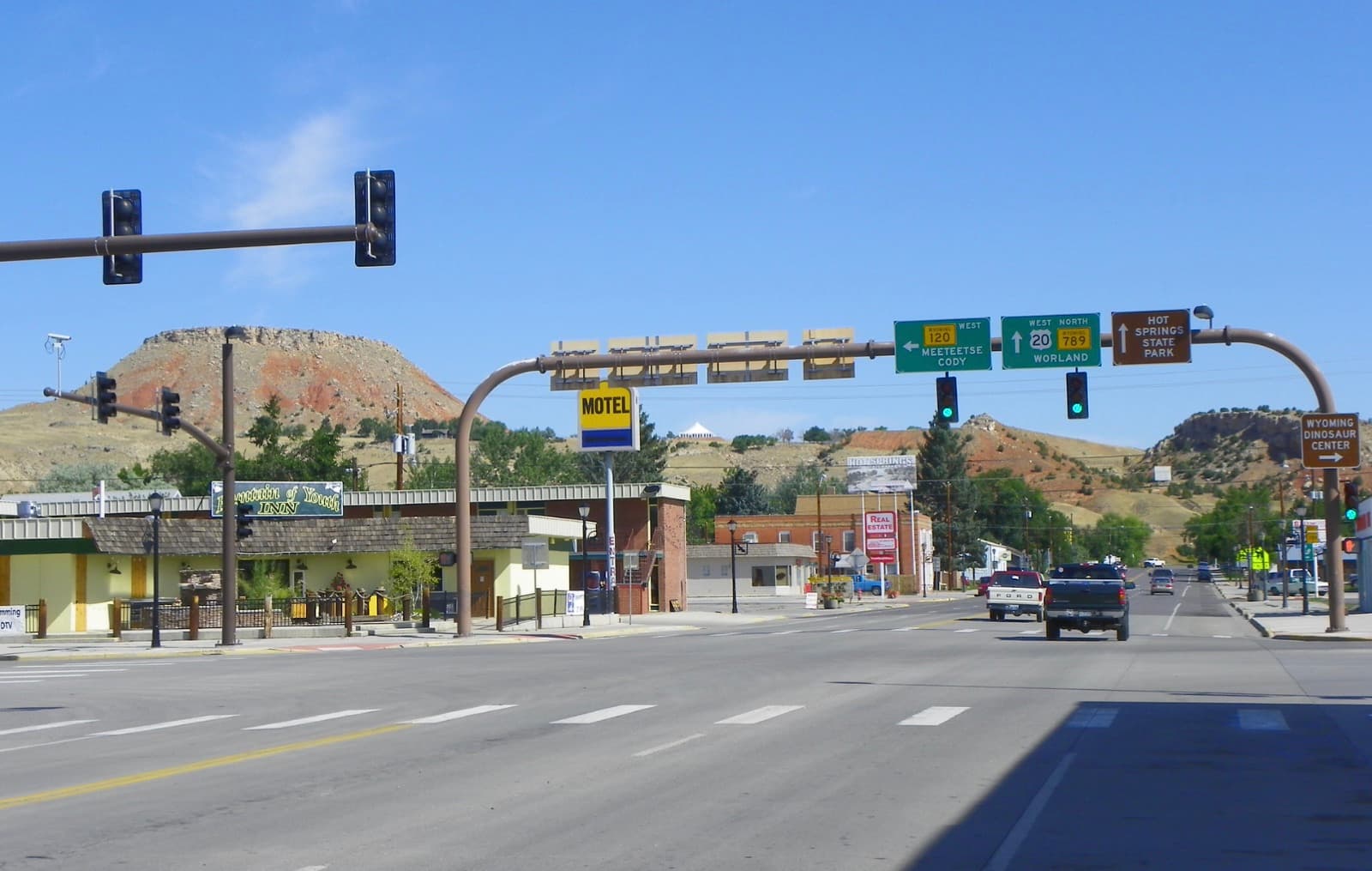 Downtown Thermopolis, Wyoming at the U.S. 20 junction — storefronts and highway signs toward Hot Springs State Park, with Round Top Mountain and red-rock bluffs behind town