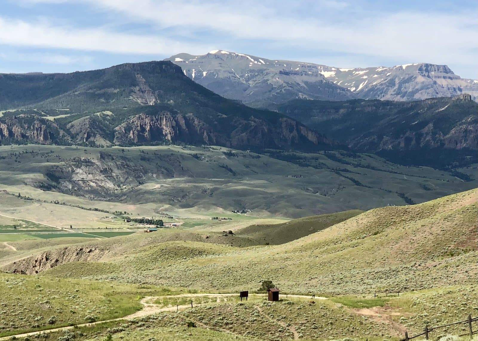 The North Fork Shoshone valley west of Cody — open ranch ground in the foreground with the snow-streaked Absaroka peaks rising along the route to Yellowstone's East Entrance